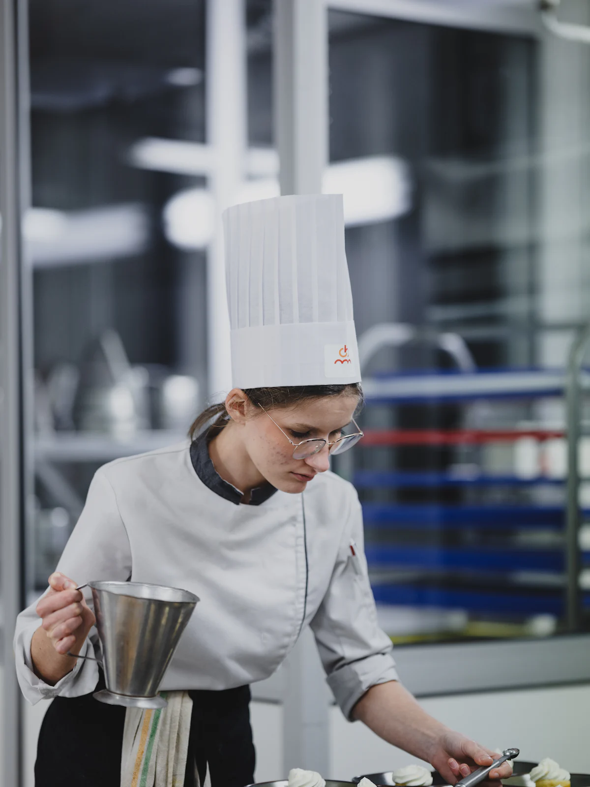 Uma aprendiz de cozinha, de chapéu alto e óculos, a finalizar pratos